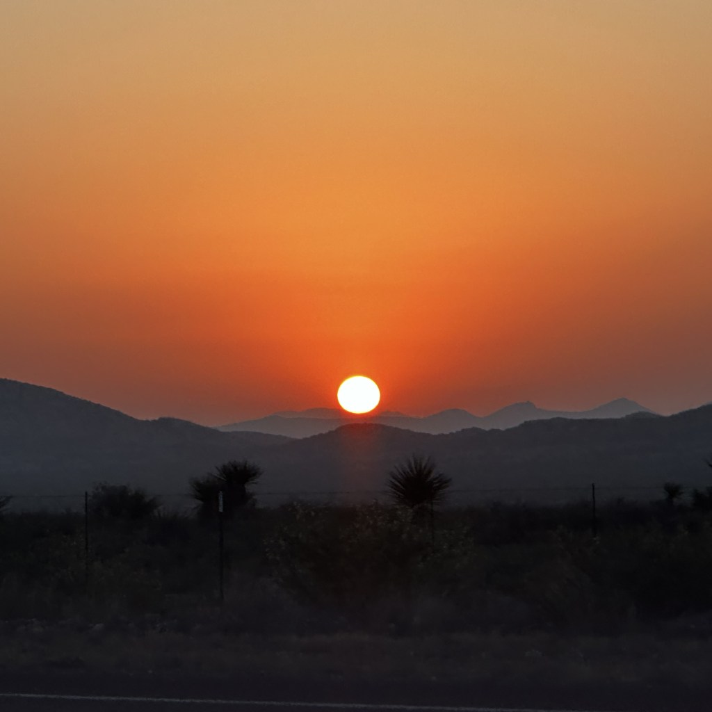 An orange sky with a bright sun just over some dark mountains.